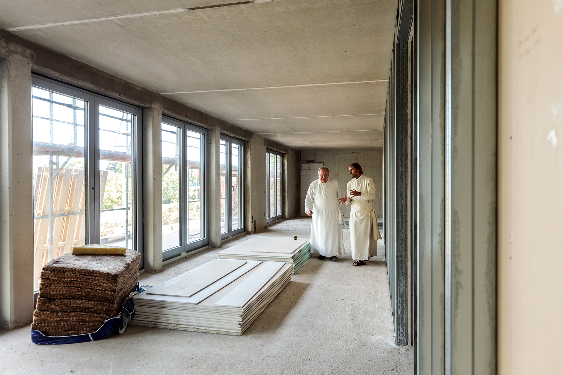 Zwei Mönche in weißen Gewändern stehen in einem unvollendeten Raum eines deutschen Klosters, umgeben von Baumaterialien und großen Fenstern.