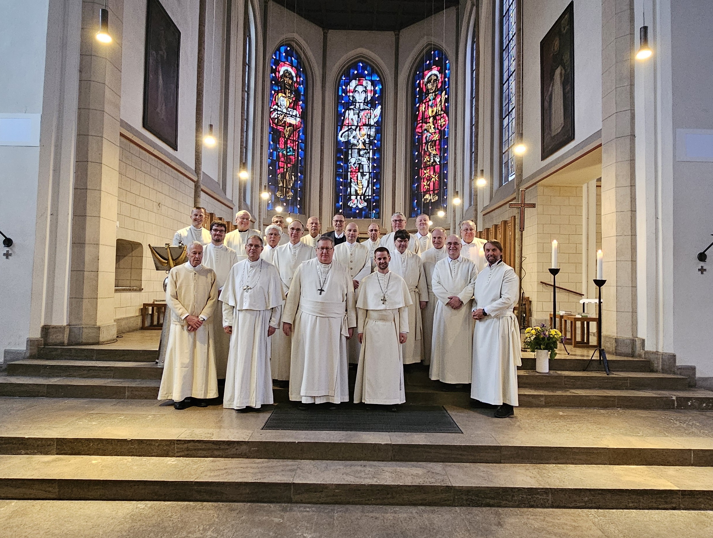 Gruppe von Geistlichen in weißen Gewändern posiert in einer Kirche mit bunten Glasfenstern – vermutlich ein Kloster in Deutschland.