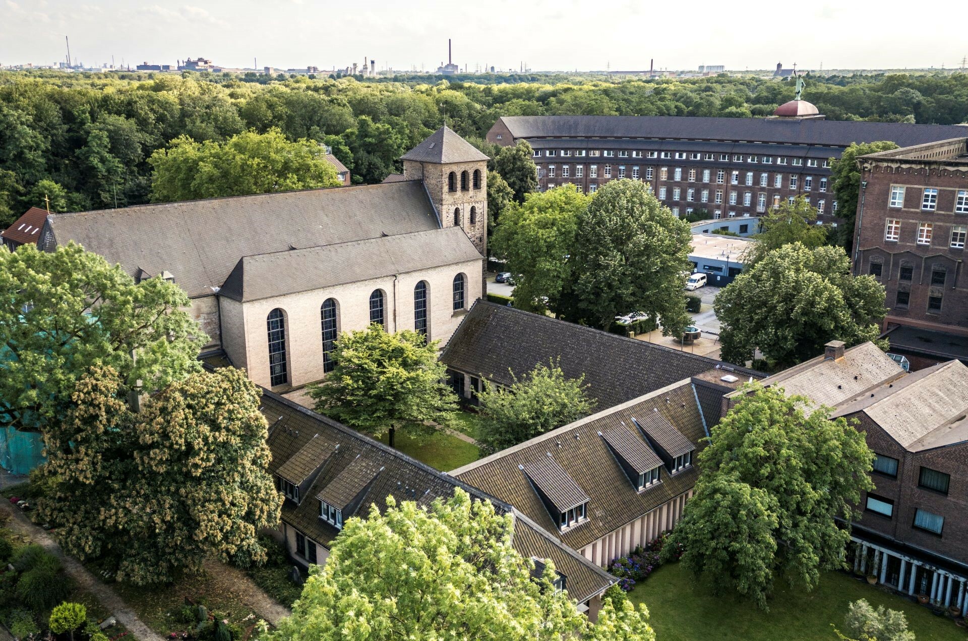Luftaufnahme eines Klosters in Deutschland: Kirche mit Turm, umgebende Gebäude, dichte Bäume und Industriefabriken im Hintergrund.