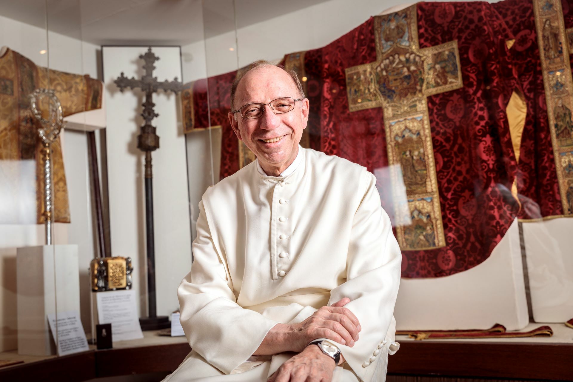 Ein Mönch in weißer liturgischer Robe sitzt vor religiösen Exponaten, darunter einem Kreuz und einem roten bestickten Messgewand, in einem deutschen Kloster.