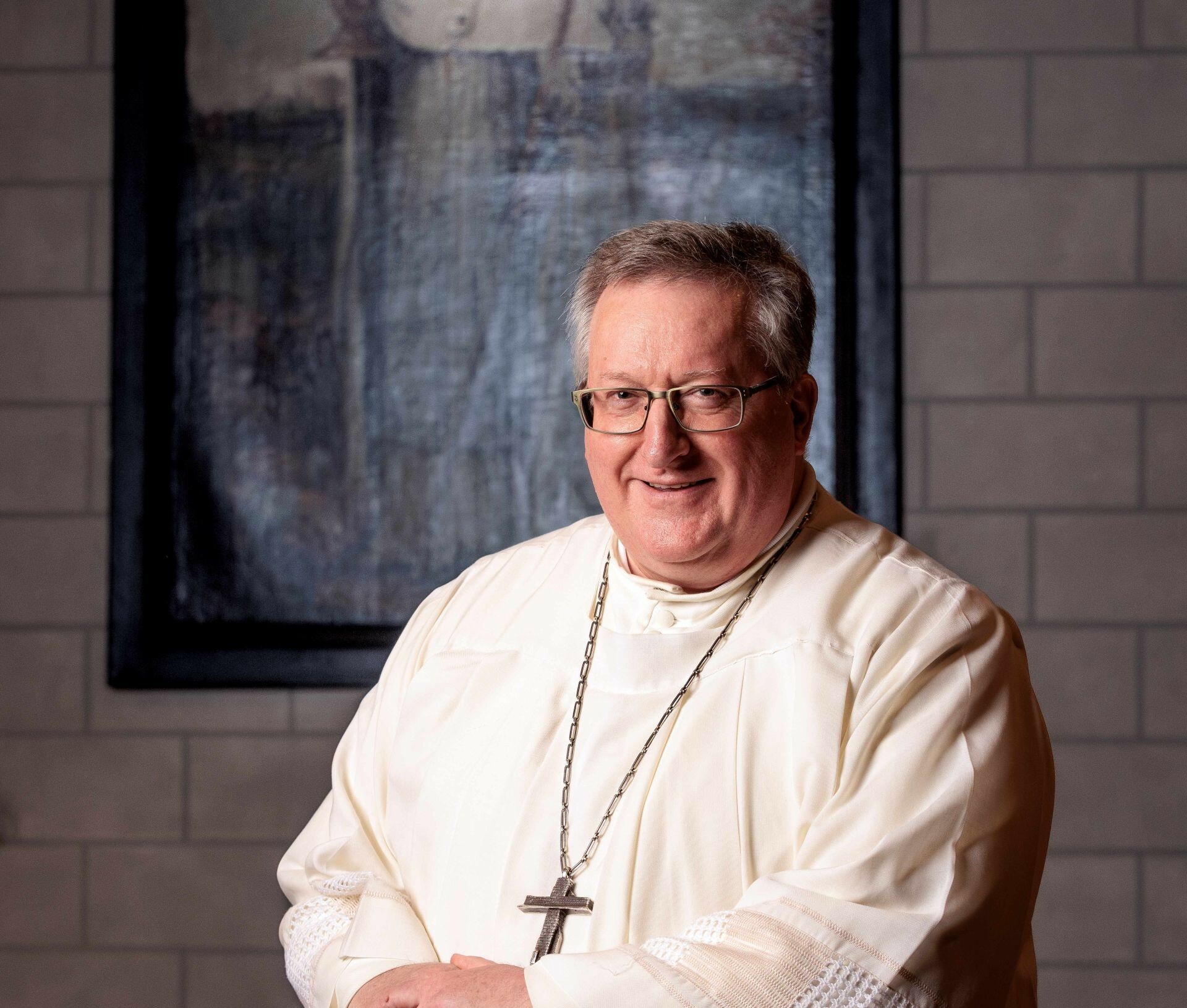 Ein Priester in weißer liturgischer Robe mit Kreuzanhänger steht vor einer Ziegelwand mit gerahmtem Gemälde im Hintergrund, in einem deutschen Kloster.